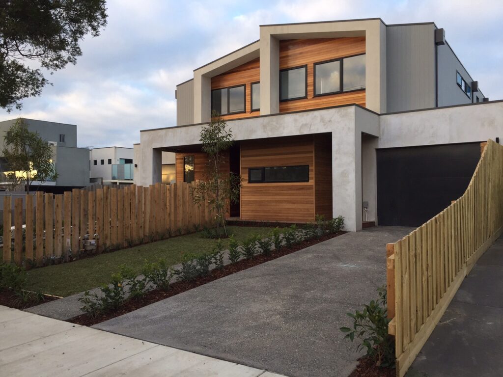 Modern two-story house with a wooden accent facade, large windows, and a black garage door. Surrounded by a garden and wooden fence. Cloudy sky above.