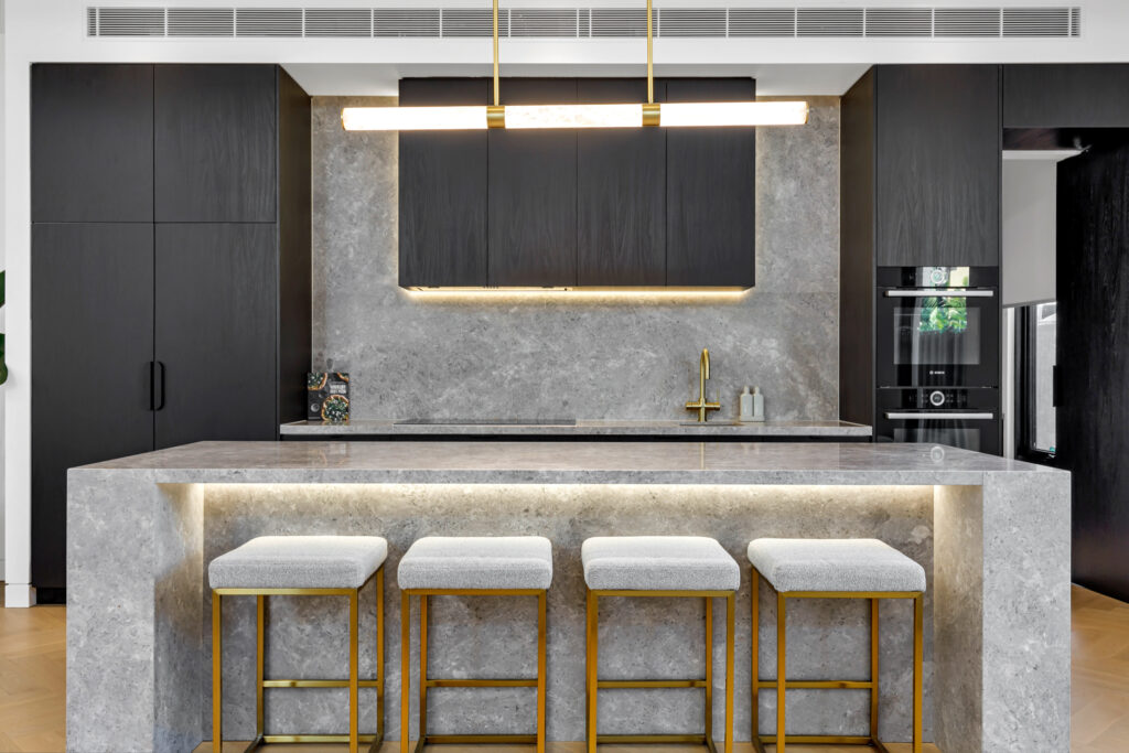 Modern kitchen with a sleek, grey stone island and four cushioned stools. Dark cabinets contrast with golden faucet and under-cabinet lighting. Stylish ambiance.