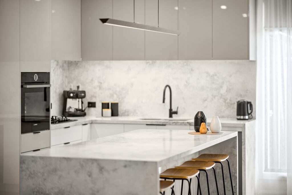Modern kitchen with sleek white marble countertops, grey cabinets, and a black faucet. Features a pendant light, barstools, and minimalist decor.
